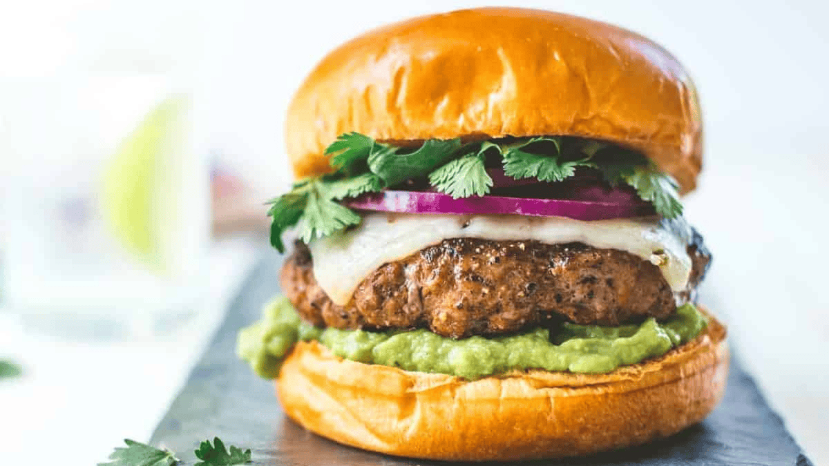 A juicy burger with melted cheese, red onion, fresh cilantro, and guacamole on a soft golden bun, served on a dark slate board.