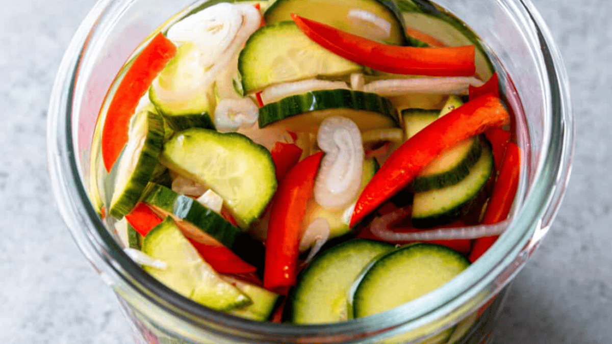 A glass jar filled with pickled vegetables, including sliced cucumbers, red bell peppers, and onions, in a light brine. The colorful ingredients are visible through the clear jar.