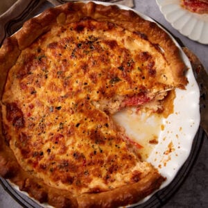 A baked savory pimento cheese tomato pie with a golden, cheesy crust sits in a white pie dish. A large slice has been removed, revealing a creamy, tomato-filled interior with crumbs and filling where the slice was taken.