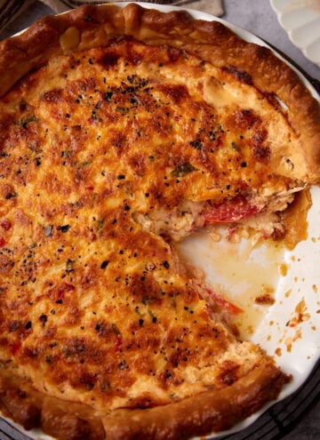 A baked savory pimento cheese tomato pie with a golden, cheesy crust sits in a white pie dish. A large slice has been removed, revealing a creamy, tomato-filled interior with crumbs and filling where the slice was taken.
