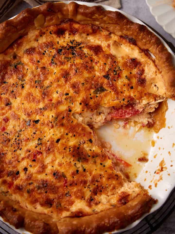 A baked savory pimento cheese tomato pie with a golden, cheesy crust sits in a white pie dish. A large slice has been removed, revealing a creamy, tomato-filled interior with crumbs and filling where the slice was taken.