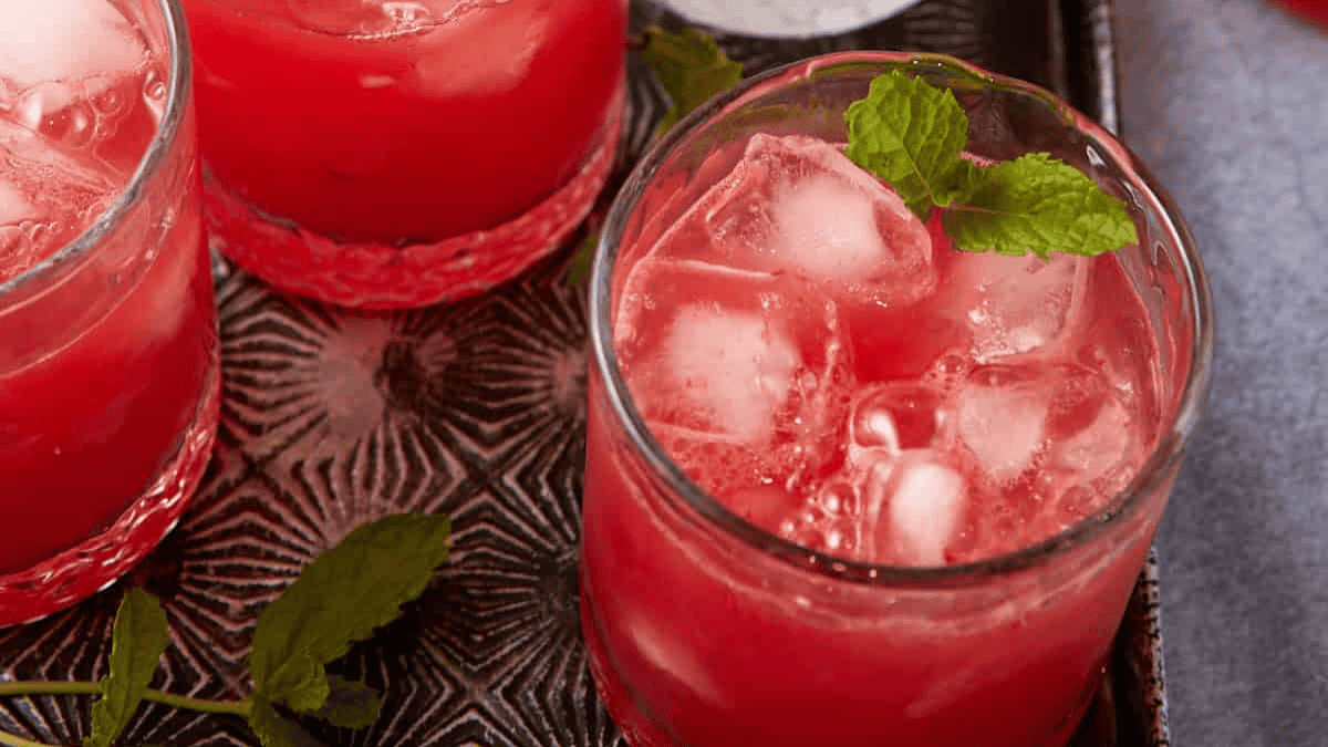 A glass of bright red watermelon drink with ice cubes, garnished with fresh mint leaves, sits on a patterned tray. Another glass and scattered mint leaves are visible nearby.