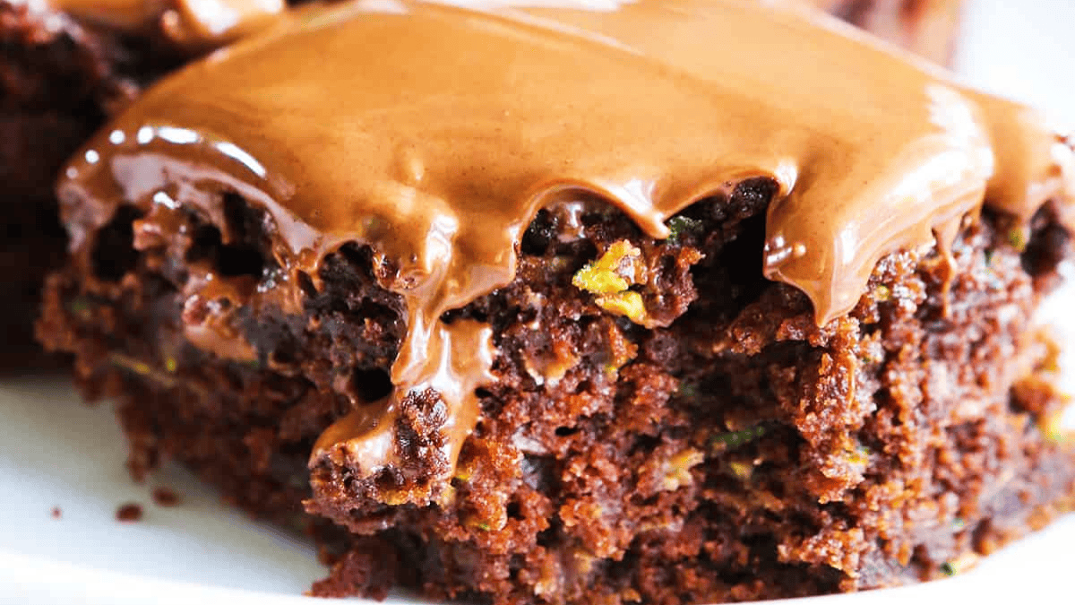 A close-up of a moist chocolate zucchini cake topped with a thick, glossy layer of melted chocolate frosting. The cake’s texture and flecks of green zucchini are visible.