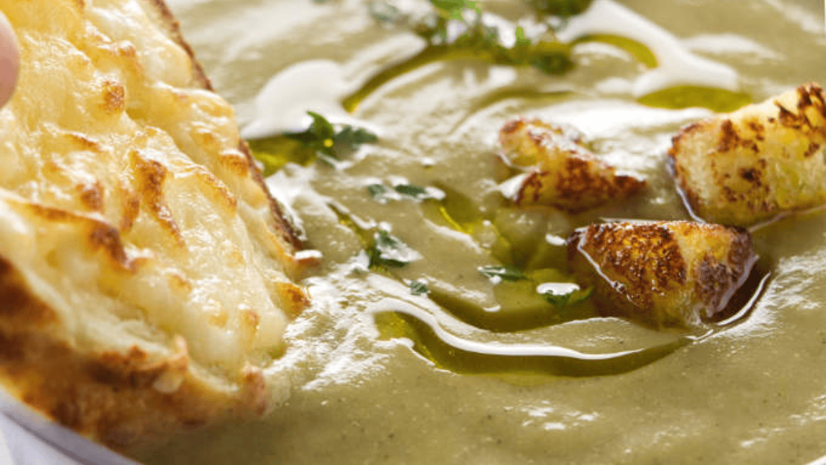 Close-up of creamy green soup topped with croutons, a swirl of olive oil, and a sprinkle of herbs, served with a piece of toasted bread covered in melted cheese.