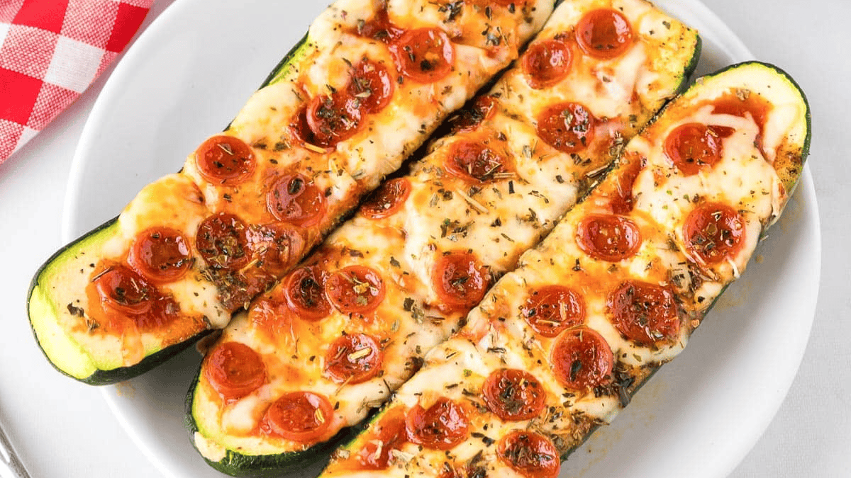 Three zucchini halves on a white plate topped with melted cheese, mini pepperoni slices, and herbs, resembling pizza boats. A red and white checkered napkin is partially visible beside the plate.