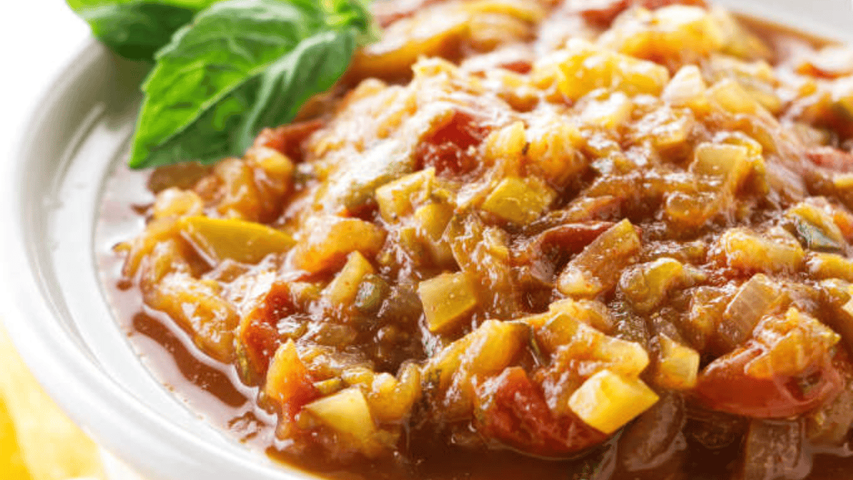 A close-up of a bowl of chunky, homemade ratatouille, featuring pieces of tomato, zucchini, and other vegetables, garnished with a fresh basil leaf.