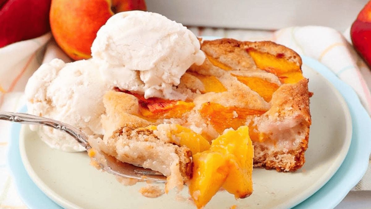 A slice of peach cobbler topped with vanilla ice cream sits on a plate, with a forkful of cobbler in the foreground. Fresh peaches are visible in the background.