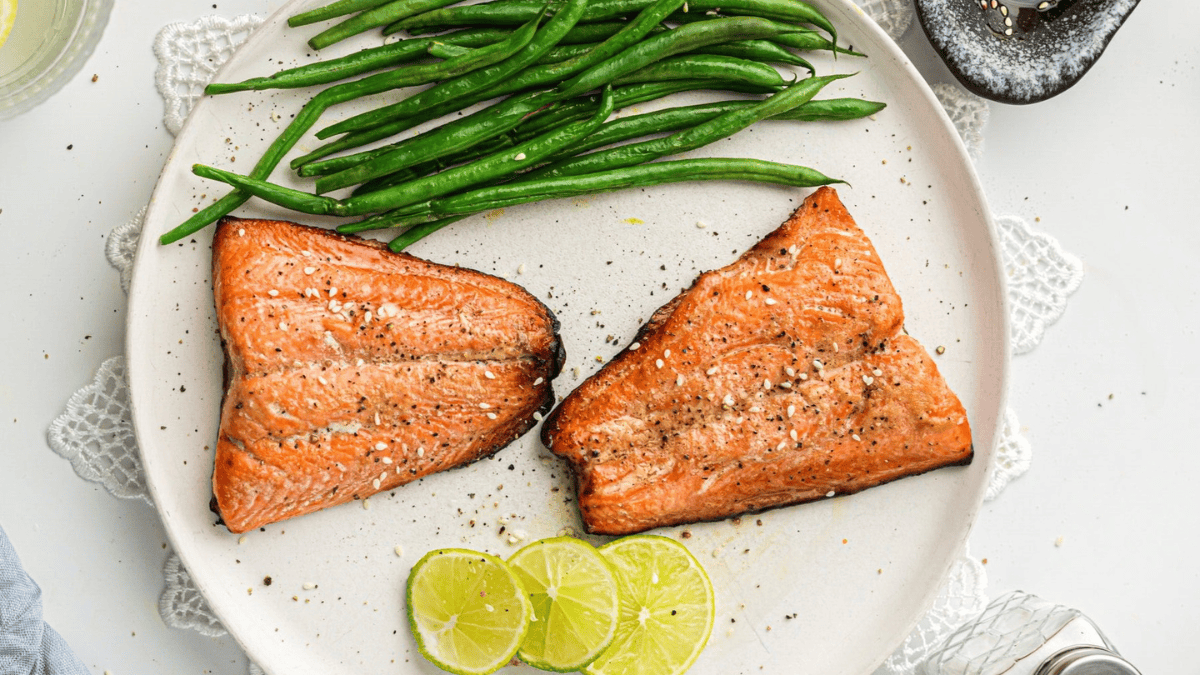 A plate with two grilled salmon fillets, fresh green beans, and three slices of lime, garnished with black pepper.