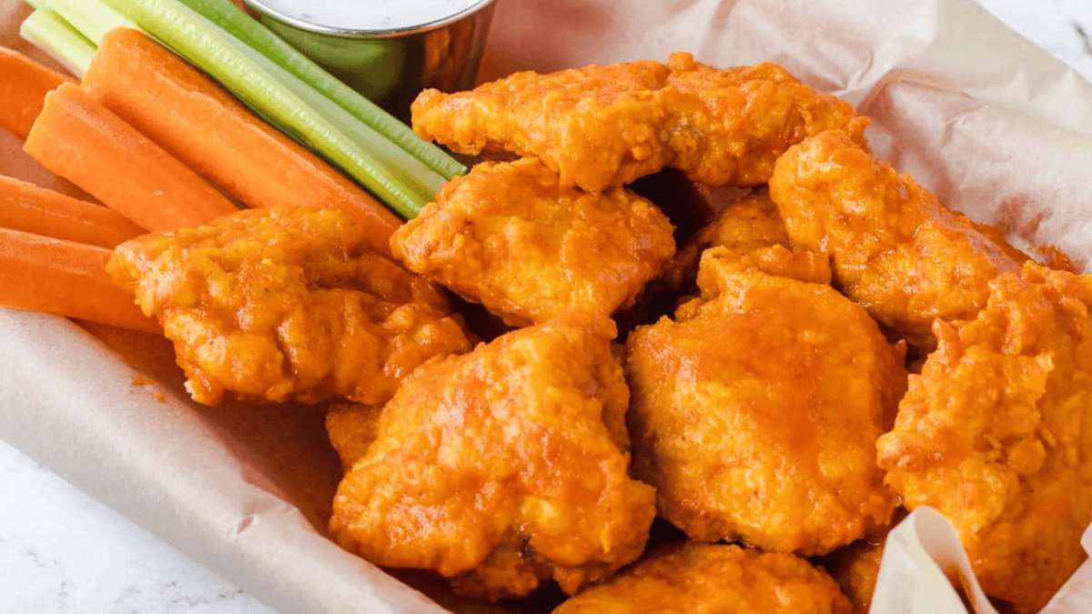 A basket lined with parchment paper holds several pieces of buffalo chicken wings, carrot and celery sticks, and a cup of ranch dipping sauce.