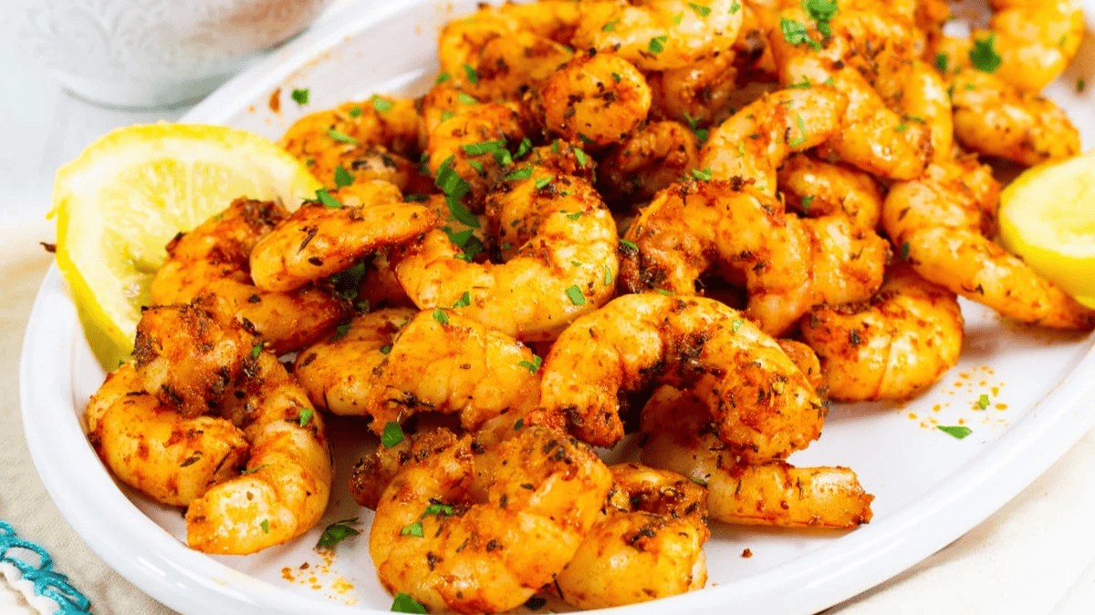 A plate of seasoned, cooked shrimp garnished with chopped parsley and lemon wedges, served on a white dish. The shrimp are coated with spices, giving them a vibrant orange color.