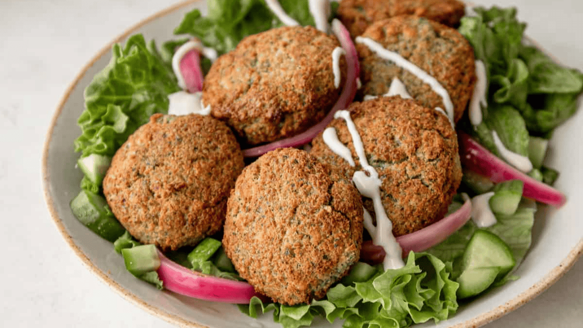 Four crispy falafel patties served on a bed of leafy greens, sliced cucumbers, and pickled red onions, drizzled with a light white sauce on a white plate.