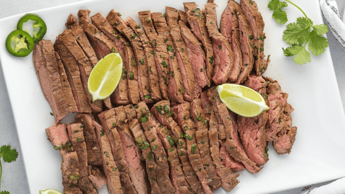 Sliced grilled steak arranged on a white platter, garnished with chopped herbs and served with lime wedges. A few jalapeño and cilantro leaves are also visible on the plate and around the edges.