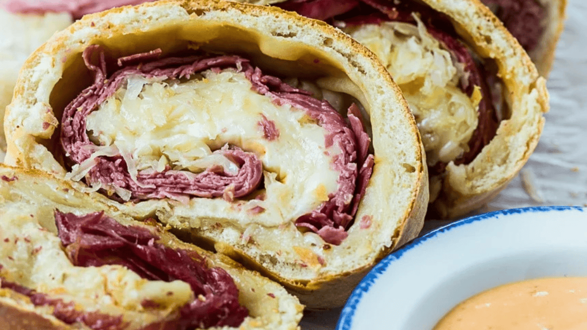 Close-up of sliced Reuben roll, showing layers of corned beef, sauerkraut, and melted cheese inside baked bread, next to a small bowl of creamy dipping sauce.