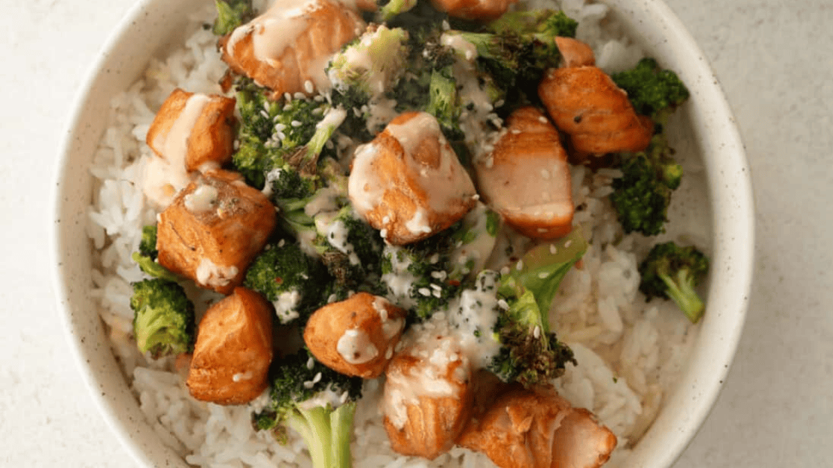 A bowl of white rice topped with cooked salmon chunks, roasted broccoli, and drizzled with a creamy sauce.
