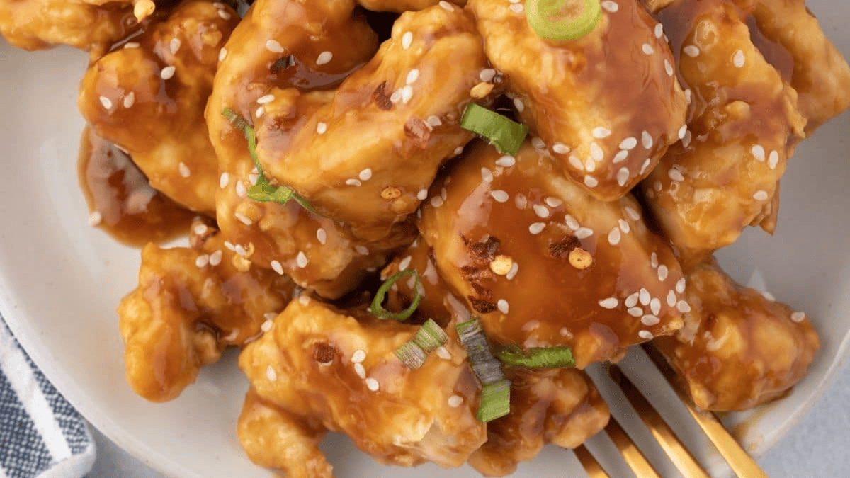 Close-up of crispy chicken pieces coated in a glossy brown sauce, topped with sesame seeds and sliced green onions, served on a white plate with a gold fork.