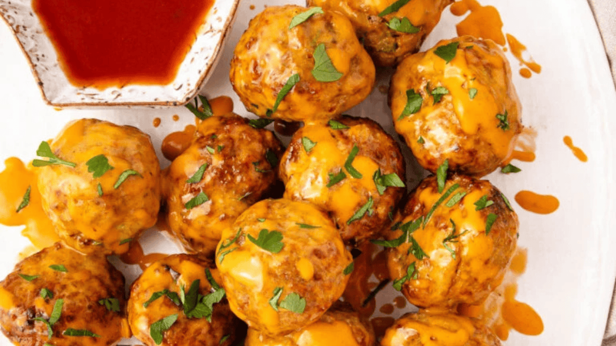 A plate of meatballs topped with melted cheddar cheese and chopped parsley, served with a side of red dipping sauce.
