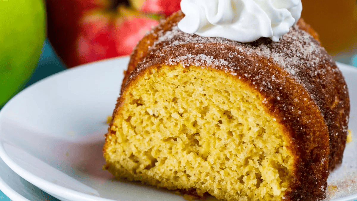 A slice of moist bundt cake dusted with sugar and topped with whipped cream sits on a white plate, with apples visible in the background.
