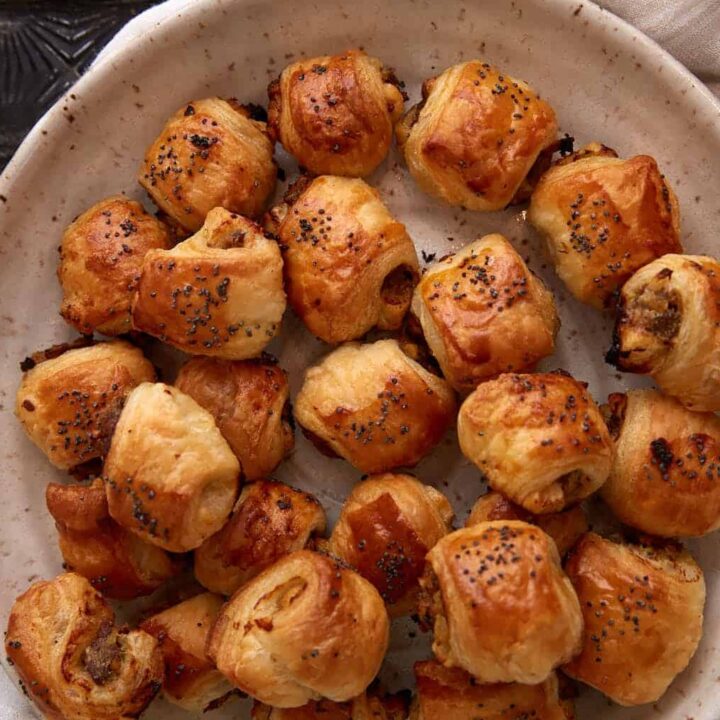 A plate filled with golden-brown sausage rolls topped with poppy seeds, arranged on a white cloth napkin. A small bowl of yellow dipping sauce is visible in the corner.