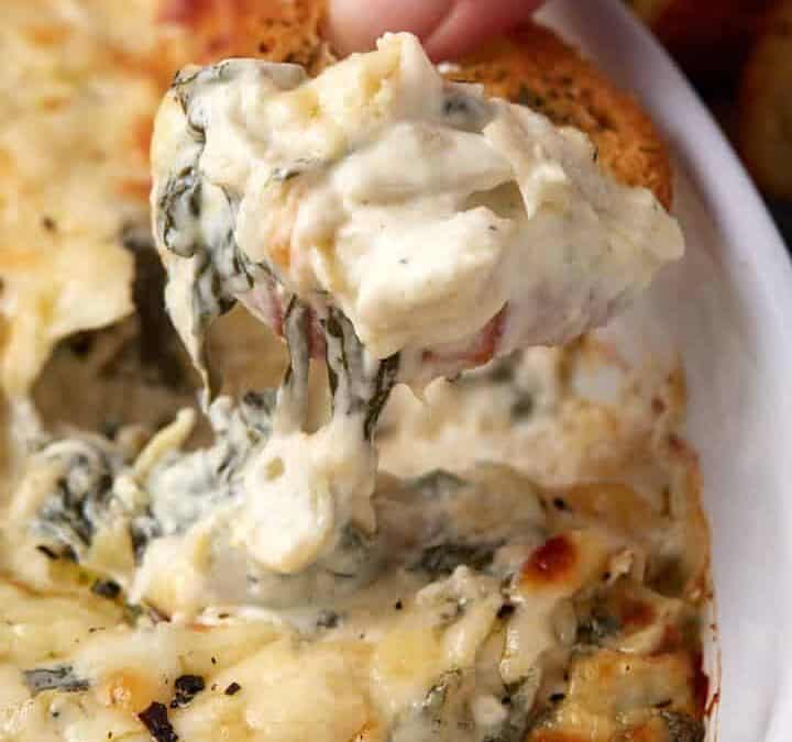 A hand dips a piece of bread into an easy spinach artichoke dip in a white casserole dish. The dip is creamy with a golden, slightly browned top—perfect for sharing at any gathering.