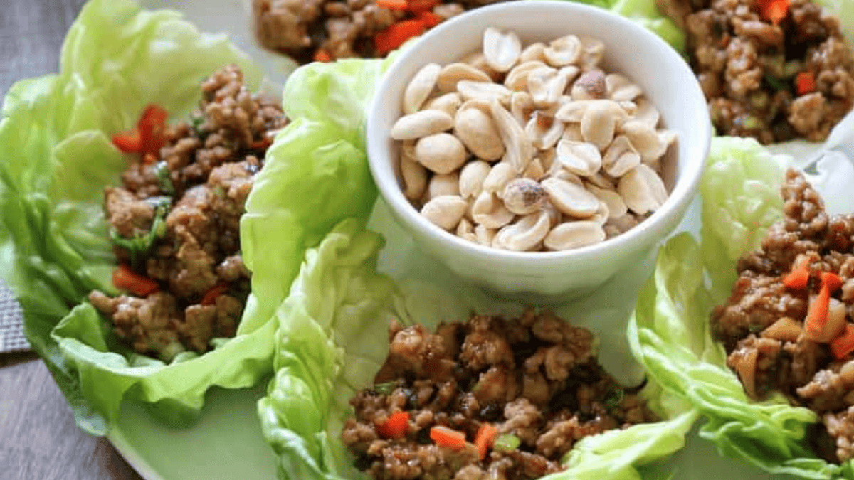 Lettuce wraps filled with minced meat and diced vegetables surround a small bowl of shelled peanuts on a green plate.