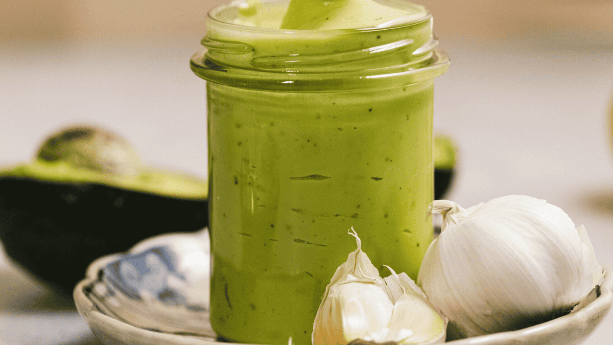 A glass jar filled with creamy green avocado sauce sits on a dish beside whole and peeled garlic cloves, with a halved avocado in the background.