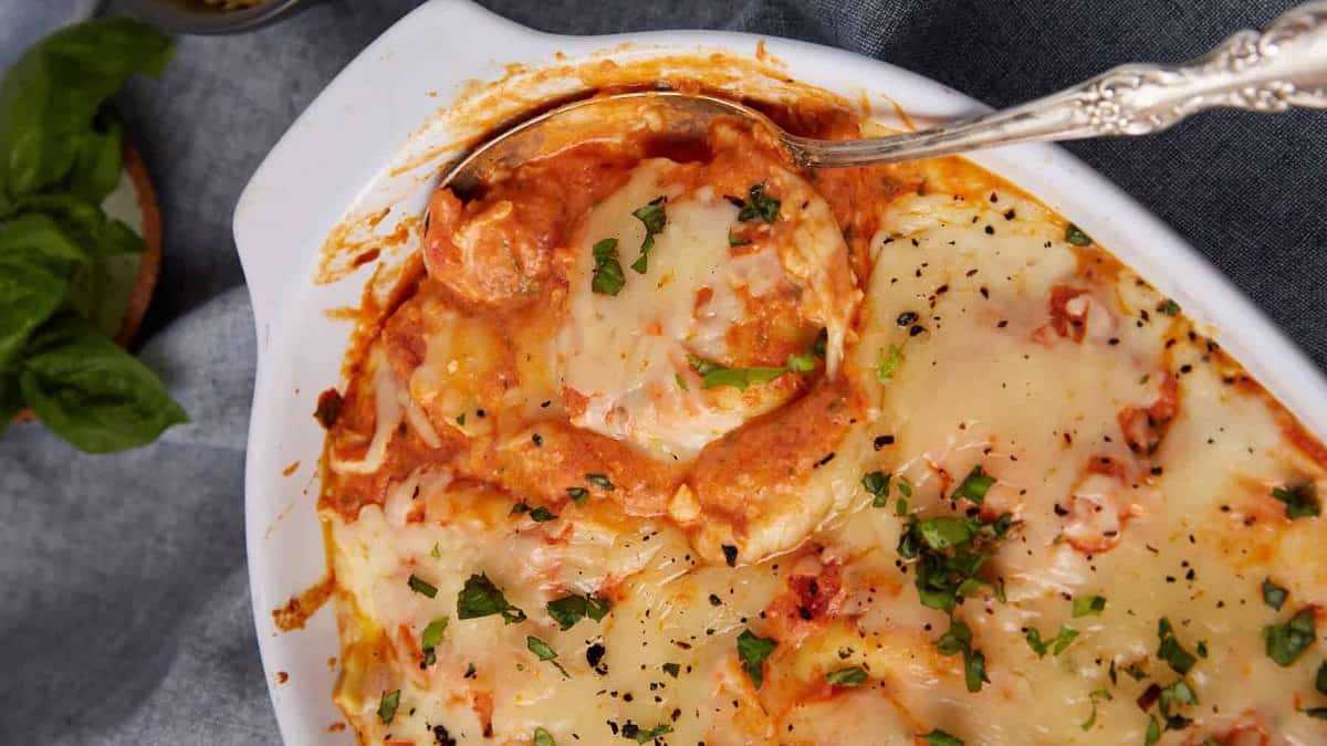 A baked dish of cheesy, saucy lasagna or Baked Ravioli with melted cheese and herbs on top, served in a white casserole dish with a silver serving spoon, next to a bowl of grated cheese and fresh basil.