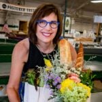 Barbara holds a tote bag filled with flowers, baguettes, and fresh produce at an indoor market.