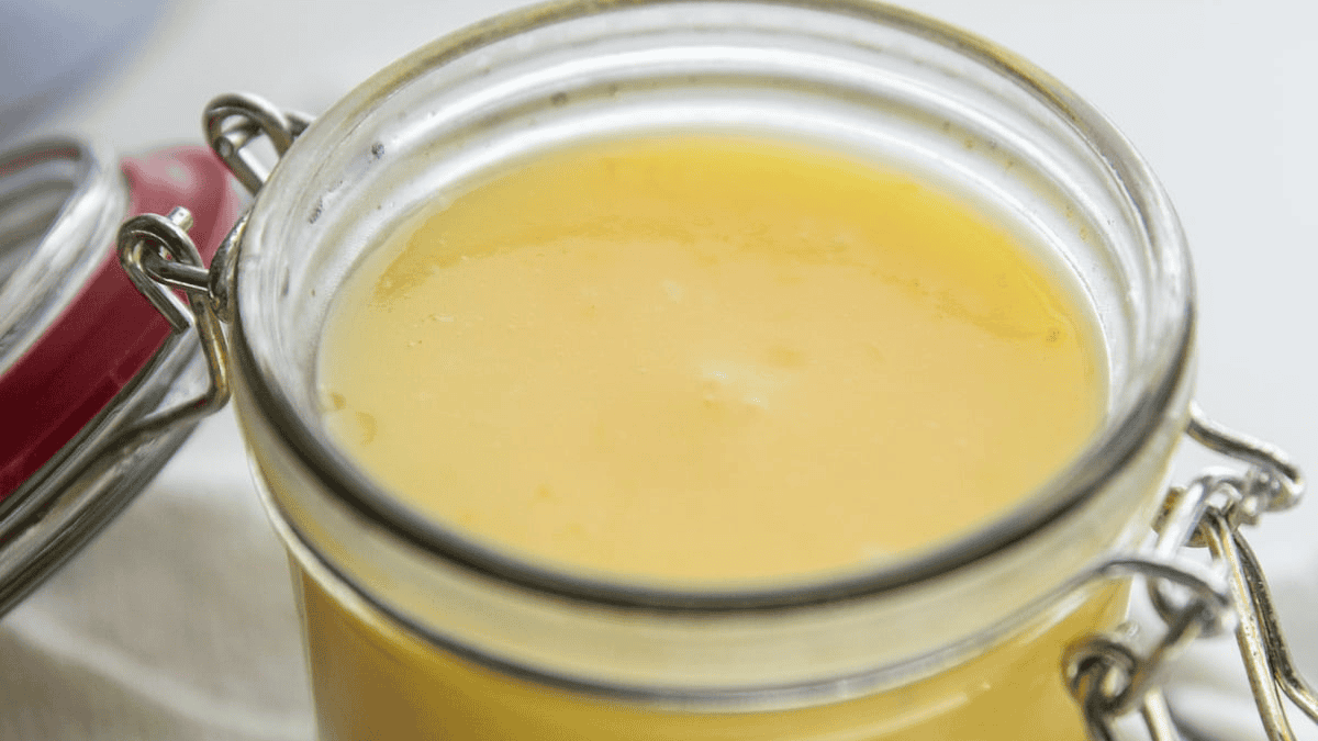 A close-up of a glass jar filled with yellow ghee, showing a smooth, glossy surface and a metal clasp lid, partially opened.