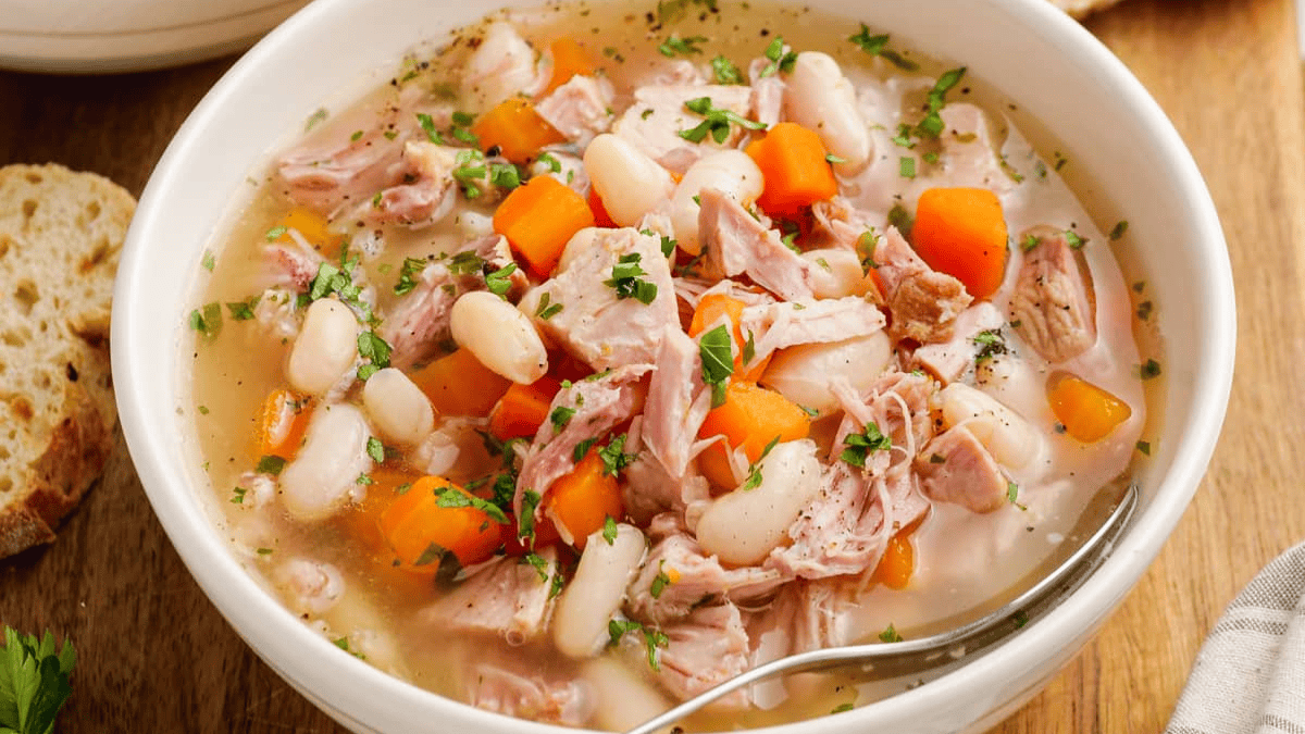 A bowl of hearty soup with chunks of ham, white beans, carrots, and fresh parsley. A spoon rests in the bowl, and slices of bread are visible on the wooden table beside it.
