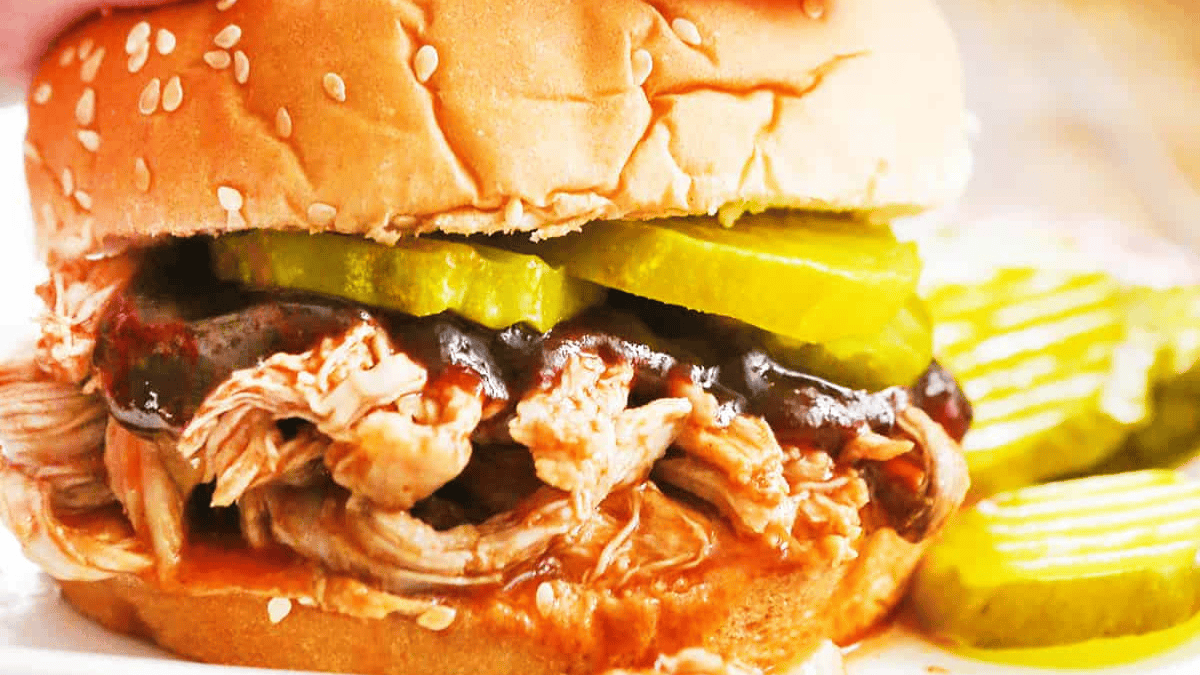 A close-up of a pulled chicken sandwich on a sesame seed bun, topped with barbecue sauce and sliced pickles. Extra pickle slices are on the side.