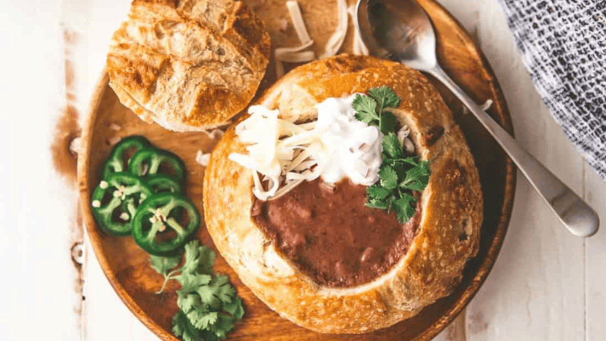 A round bread bowl filled with chili, topped with shredded cheese, sour cream, and cilantro sits on a wooden plate. Sliced jalapeños and extra cilantro are on the side, with a metal spoon next to the bowl.