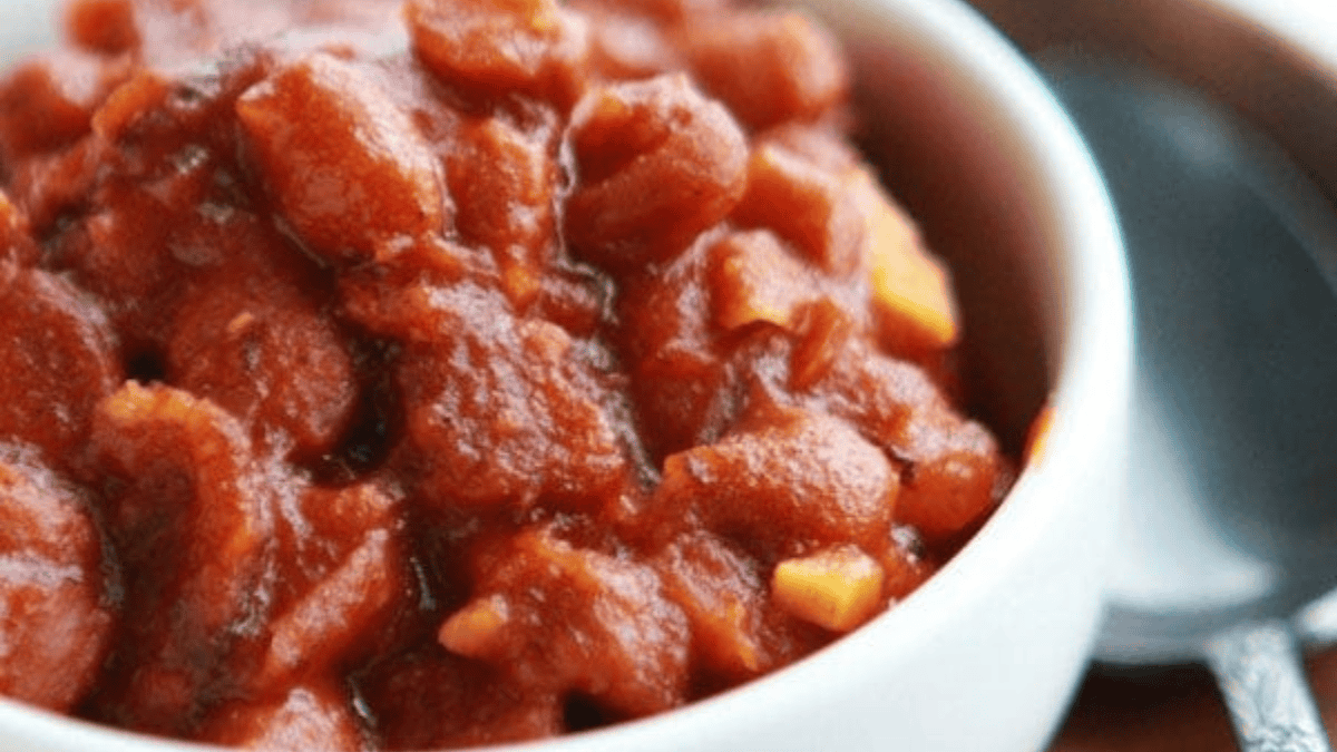 A close-up of a bowl filled with baked beans in a rich, reddish-brown sauce, with visible chunks of beans and pieces of what appears to be onion or garlic; a spoon rests beside the bowl.