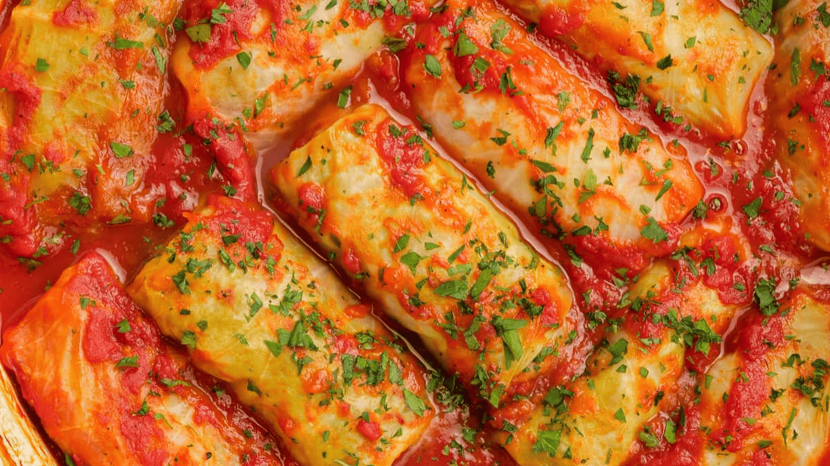 Stuffed cabbage rolls covered in tomato sauce, garnished with chopped fresh parsley, arranged closely together in a baking dish.