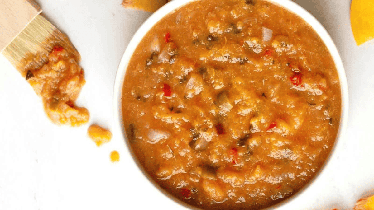 A white bowl filled with chunky, orange-brown barbecue sauce sits on a white surface, with a basting brush coated in sauce resting nearby.