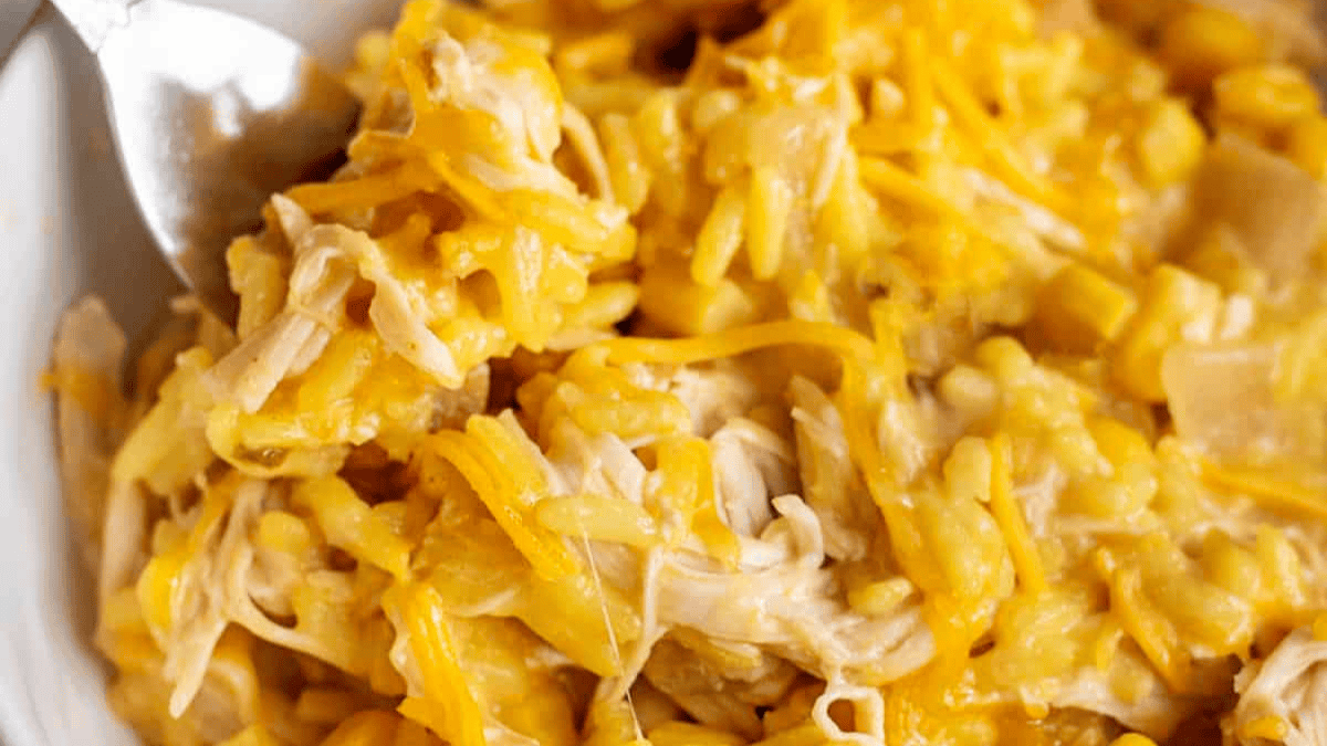 Close-up of a cheesy casserole with shredded chicken, rice, corn, and melted yellow cheese, mixed together and served in a white bowl with a fork.