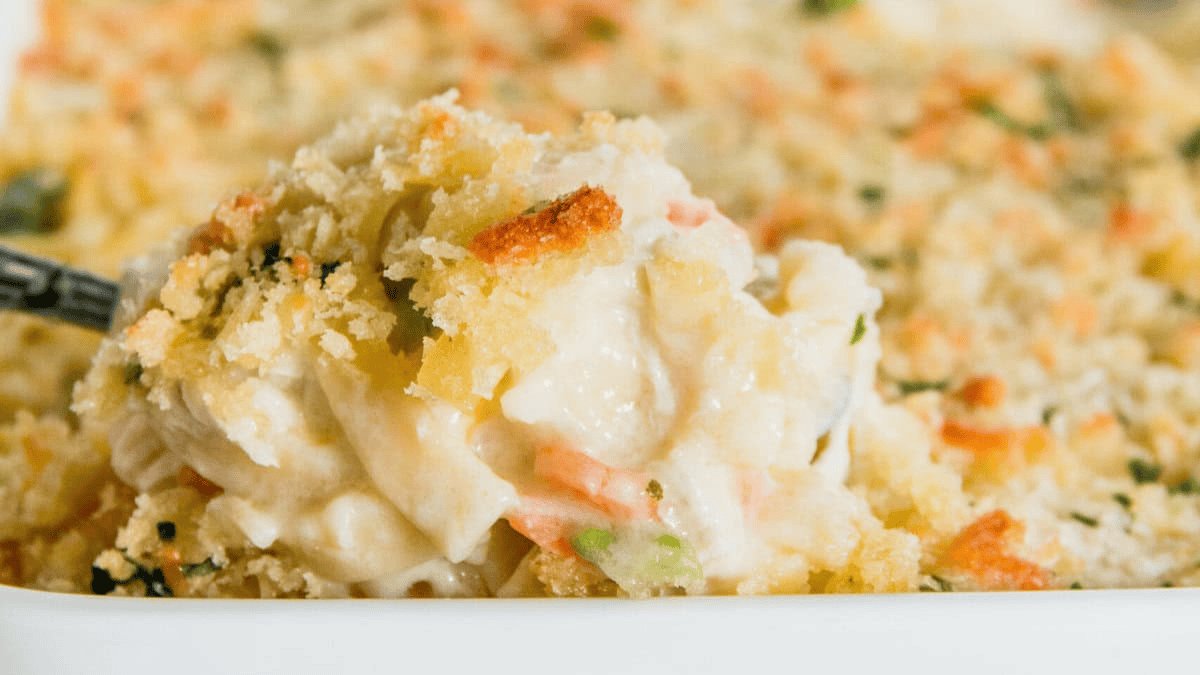 A close-up of a spoonful of creamy casserole with a golden breadcrumb topping, showing bits of vegetables like carrots and green peas in a white baking dish.