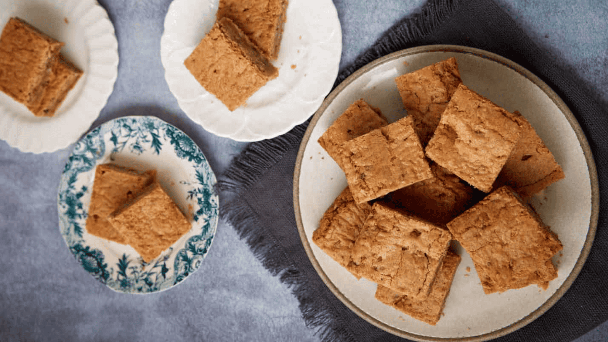 Three plates with several square blondie bars are arranged on a gray surface. One plate is white, another is white with a blue floral pattern, and the largest plate is beige and round, sitting on a dark cloth.