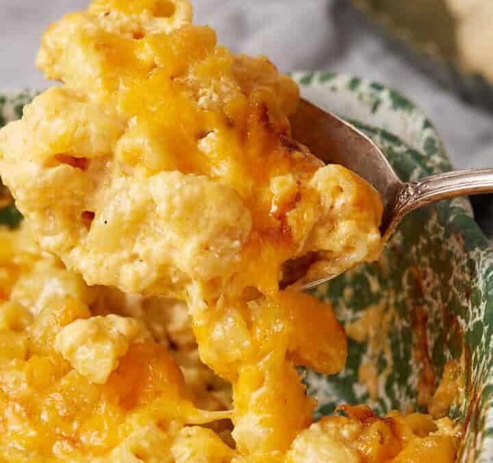 A serving spoon lifts a portion of creamy One Pot Macaroni and Cheese topped with melted cheddar from a green patterned dish, with a plate of dinner rolls in the background.