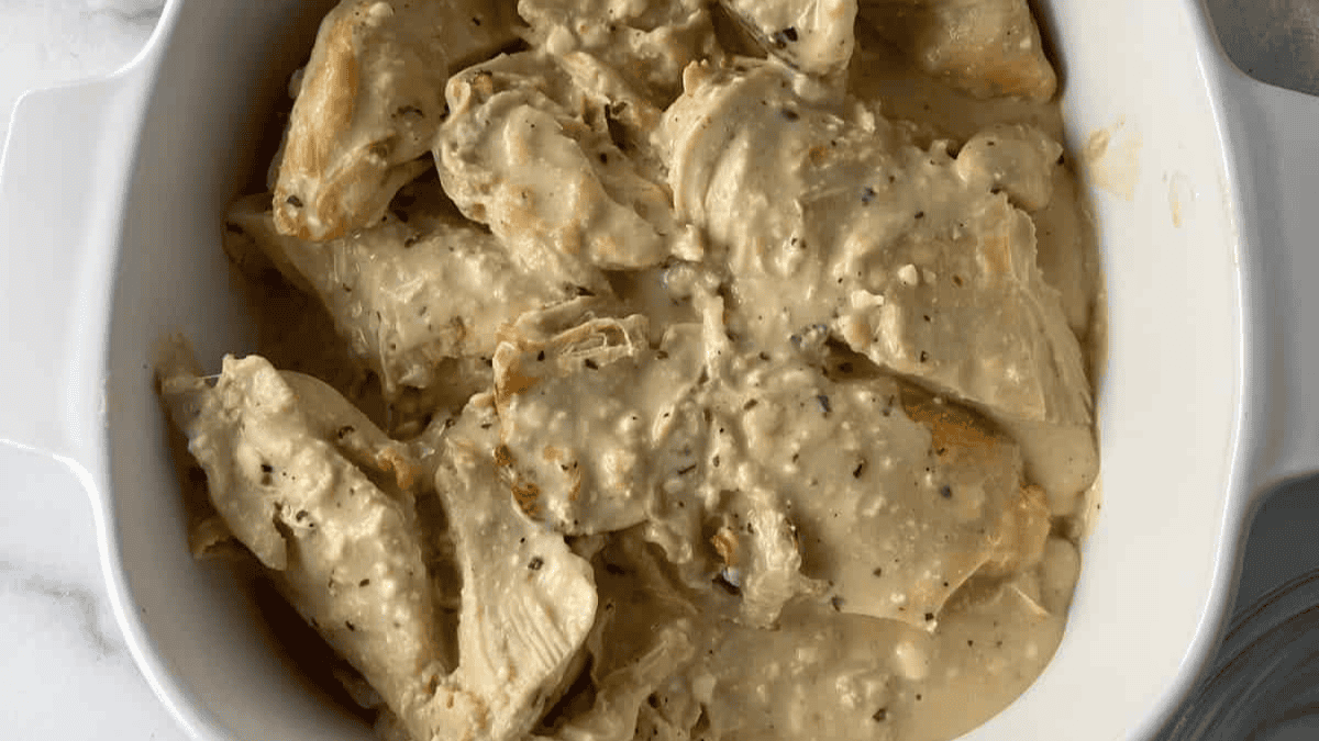 Cooked chicken breasts covered in a creamy, seasoned sauce are arranged in a white baking dish, set on a marble surface.