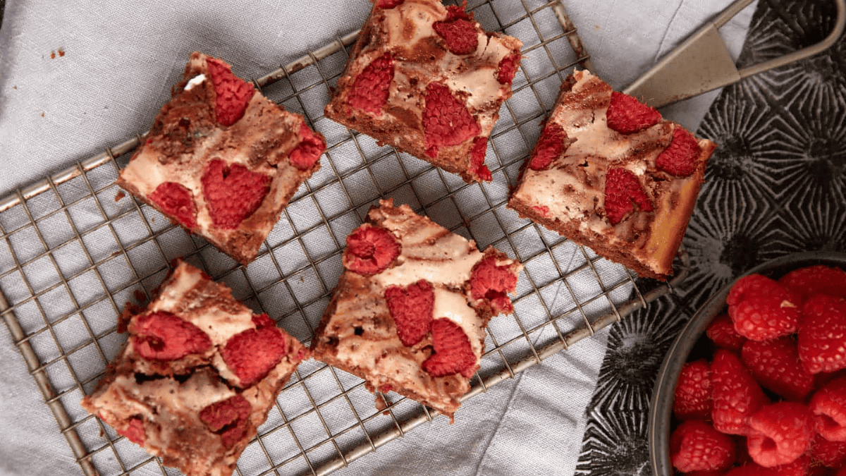 Five raspberry cheesecake brownies sit on a wire cooling rack, with a bowl of fresh raspberries and a cake server nearby. The brownies have a marbled top with baked raspberries.