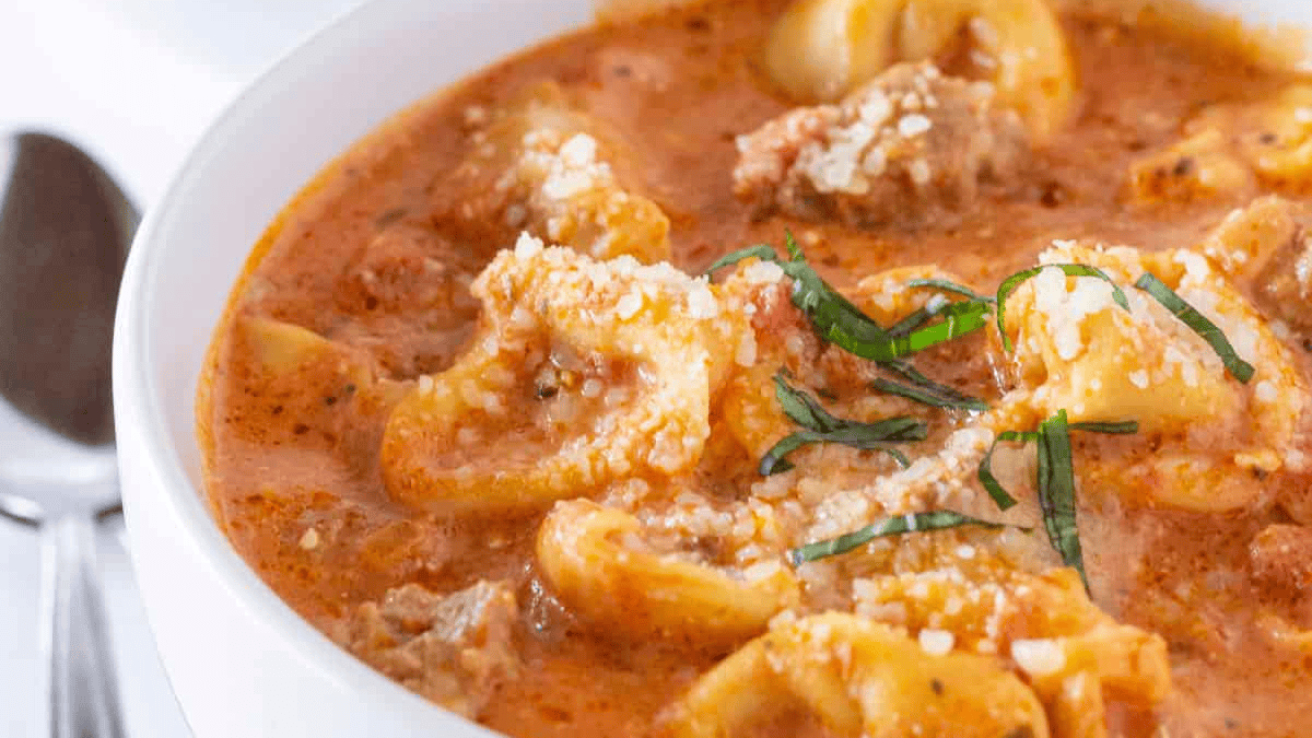 A close-up of creamy tortellini soup in a white bowl, topped with grated cheese and sliced fresh basil, with a spoon visible in the background.