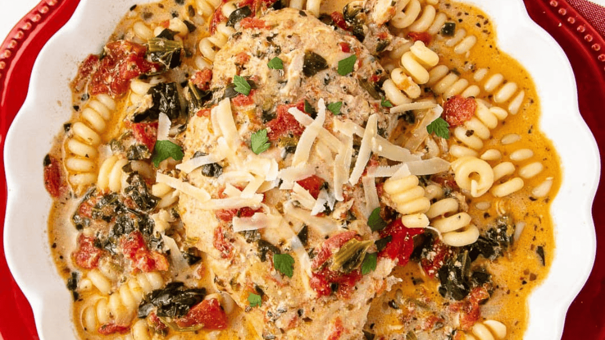 A creamy Tuscan chicken breast served in a white dish with spiral pasta, spinach, sun-dried tomatoes, and a sprinkle of shredded cheese, garnished with parsley.