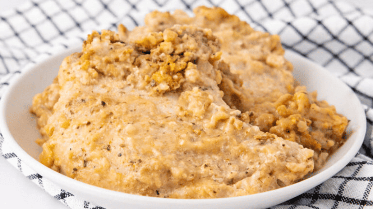A white bowl holds two creamy, golden-brown chicken breasts with stuffing, set on a white and black checkered cloth. The chicken is covered in a rich, textured sauce with visible herbs.