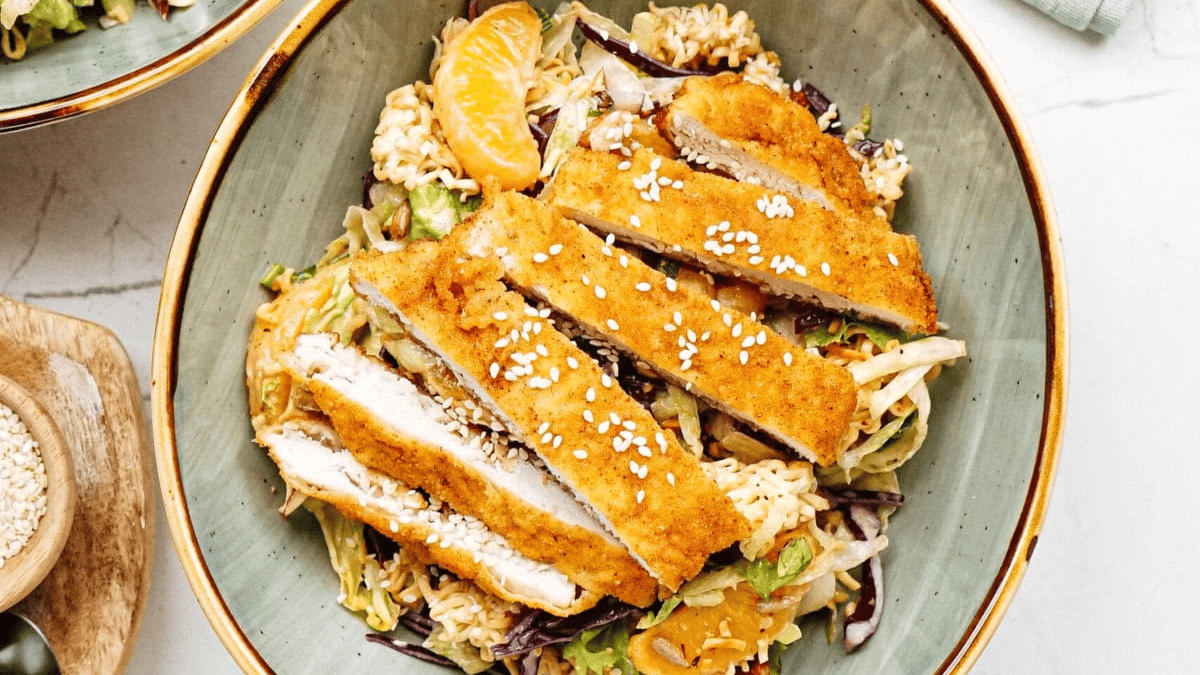 A bowl of crispy breaded chicken slices topped with sesame seeds, served over a bed of cabbage salad, noodles, and mandarin orange segments.