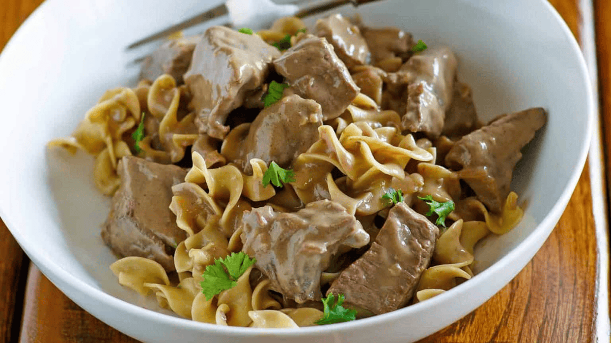 A white bowl filled with egg noodles topped with creamy beef chunks and garnished with fresh parsley, placed on a wooden surface with a fork in the background.