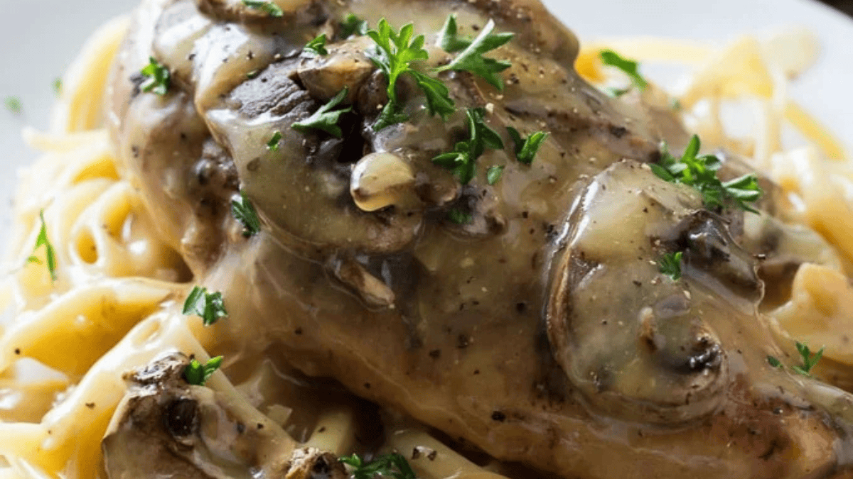 A close-up of chicken breast topped with mushrooms and creamy sauce, garnished with parsley, served over a bed of fettuccine pasta.