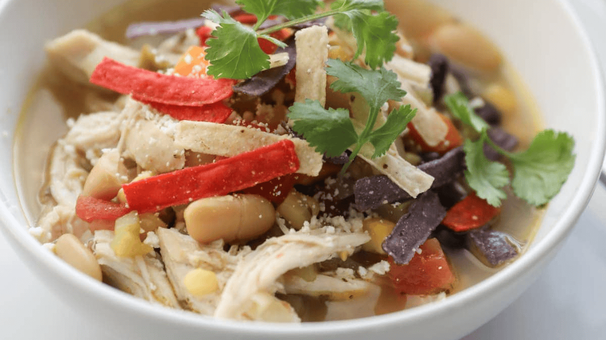 A bowl of chicken tortilla soup topped with shredded chicken, white beans, tortilla strips in red, white, and blue colors, chopped tomatoes, and fresh cilantro leaves.