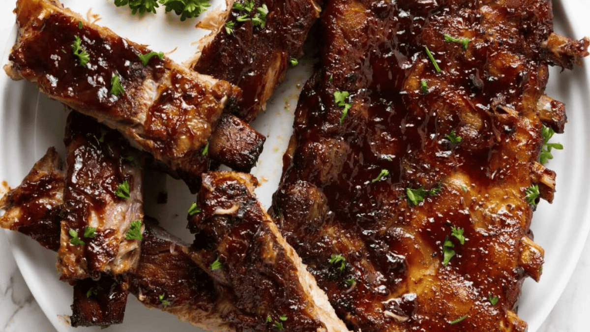 Barbecue ribs glazed with sauce, garnished with chopped parsley, served on a white plate. Some ribs are sliced, showing the juicy, tender meat.