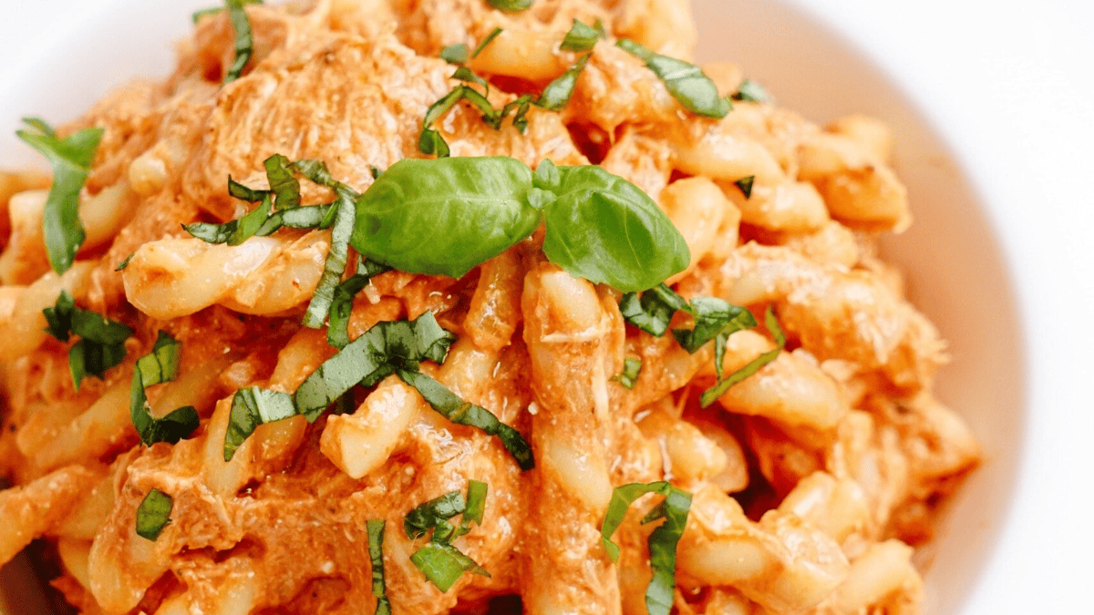 A close-up of creamy pasta with shredded chicken, tomato sauce, and fresh basil leaves, served in a white bowl.