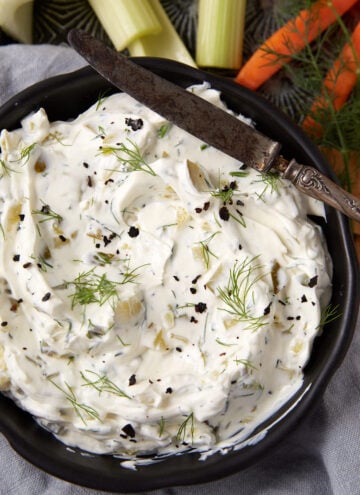 A black bowl filled with creamy dill pickle dip, garnished with fresh dill and cracked pepper; a vintage knife rests on the bowl. Celery, carrot sticks, and crackers are arranged nearby on a gray cloth.